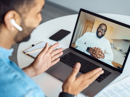 Image of a man wearing earbuds talking to a man wearing glasses via a virtual call on his computer, representing Genetx CARES Patient Navigators
